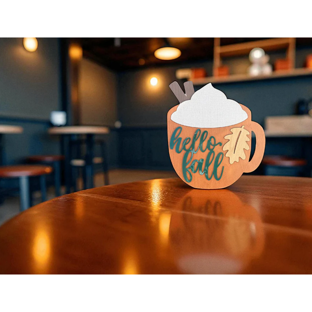 Decorative sign shaped like a coffee cup with whipped cream and chocolate pieces, featuring 'hello fall' text, on a wooden table in a cozy cafe setting. Made from Truflat cherry engineered laser plywood by Thunder Laser Canada